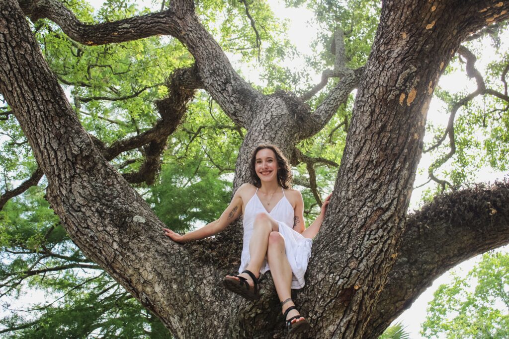 A woman in a white dress sits in a tree, with sunlight streaming down from above, creating a glowing effect in this beautiful graduation photography shot.