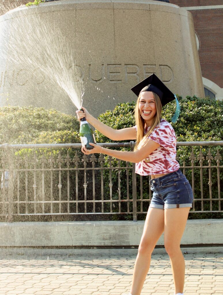 A woman in a graduation cap with a blue tassel sprays champagne in front of the Unconquered Statue at Florida State University, celebrating her graduation.