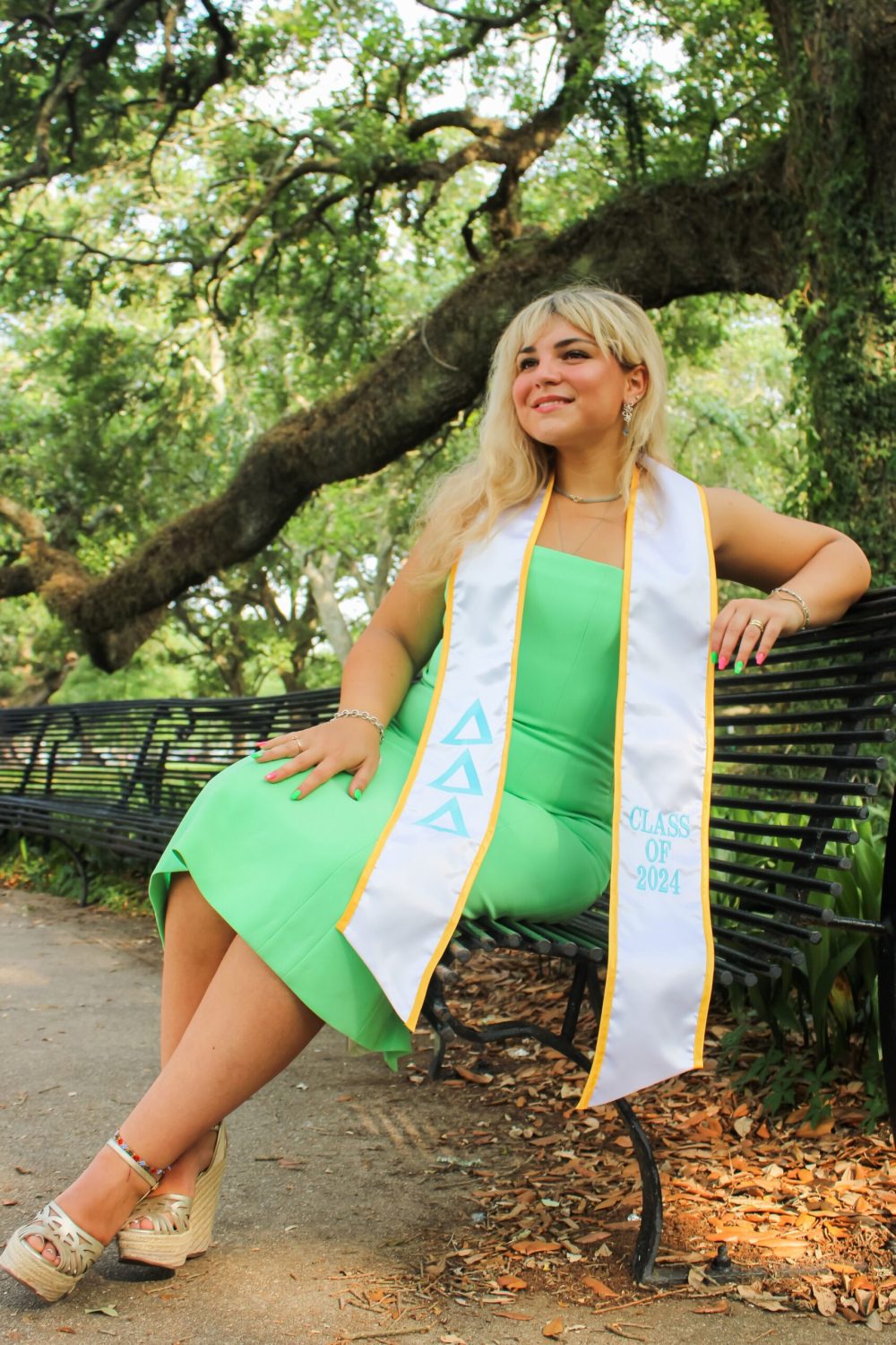 A woman in a green dress sits on a bench in front of lush foliage, wearing a white graduation tassel with yellow trim and the Greek letters 'Delta Delta Delta' stitched in light blue.