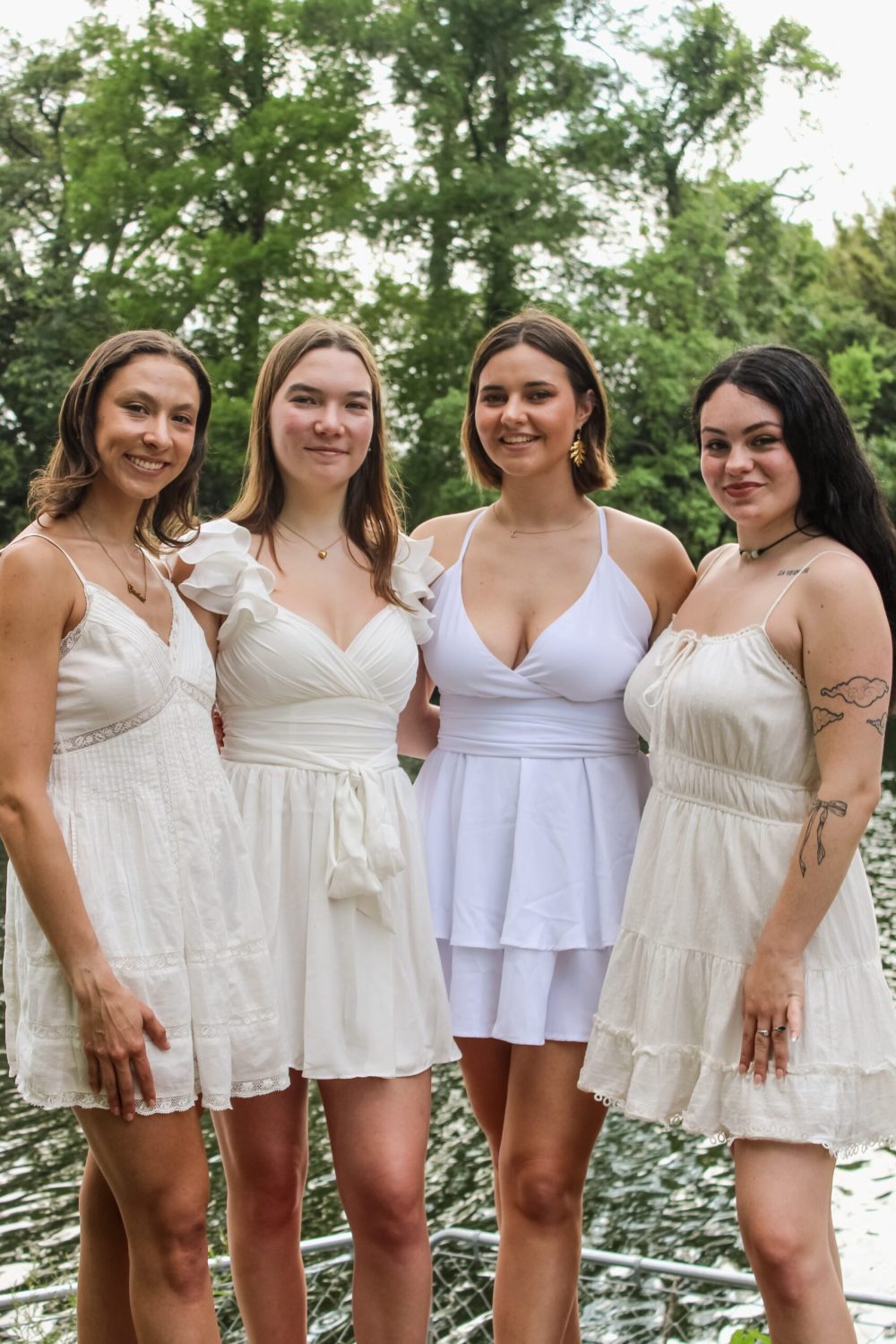 Four women in cream-colored dresses stand together in front of a lake with trees in the background, smiling and celebrating during their graduation photography session.